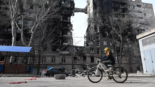 Жительница Мариуполя едет на велосипеде по улице мимо разрушенного дома